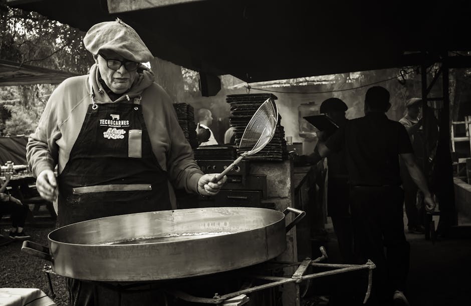 A middle-aged man cooks at an outdoor market stall using a large pan, creating street food in a lively setting.